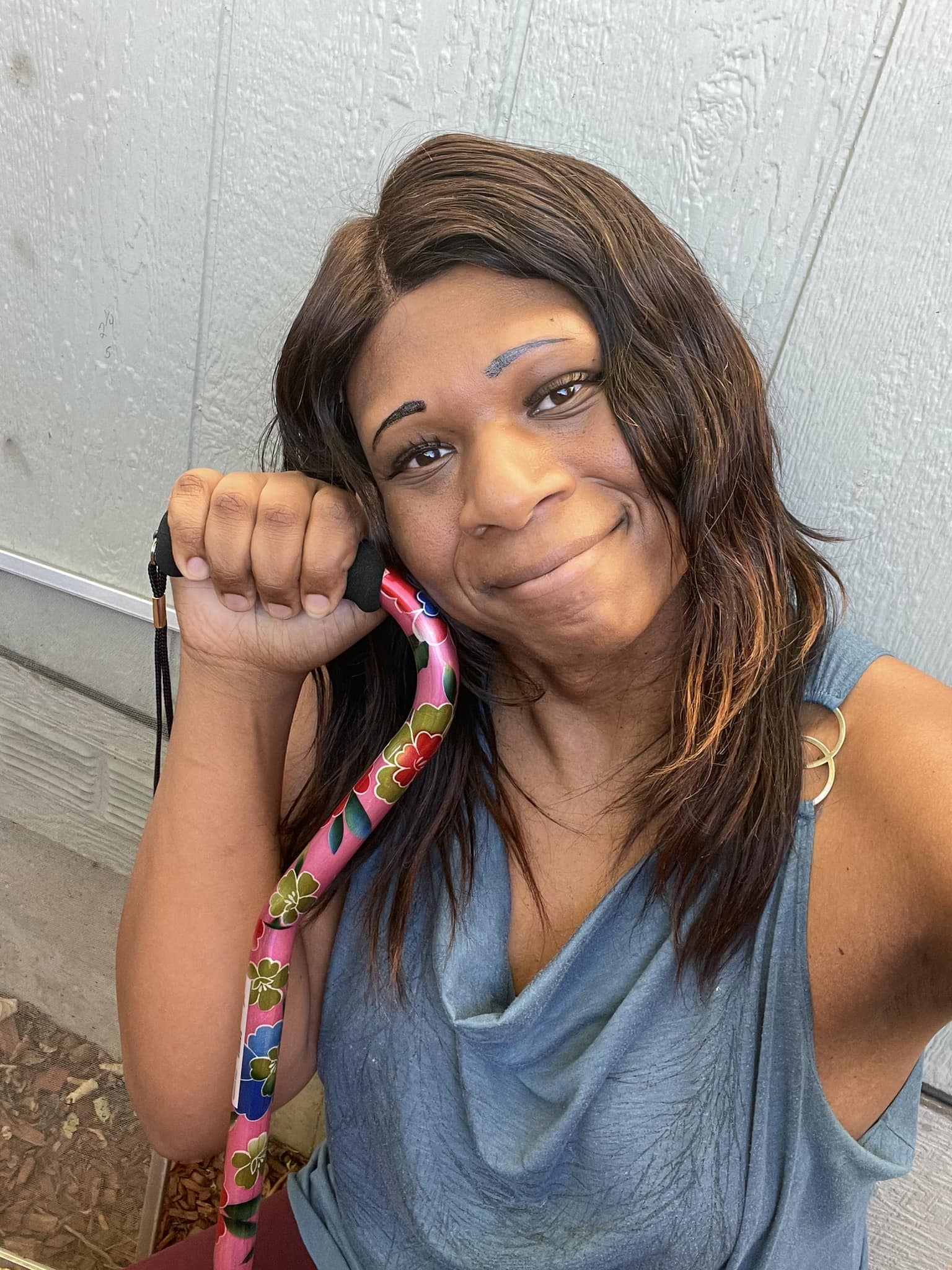 Picture of Hannah - a black trans woman crouching on a concrete porch and leaning against a green colored wall of house. She has long black hair, a teal shirt with a picture of a wolf and is smiling. She is proudly holding a cane in her right hand.